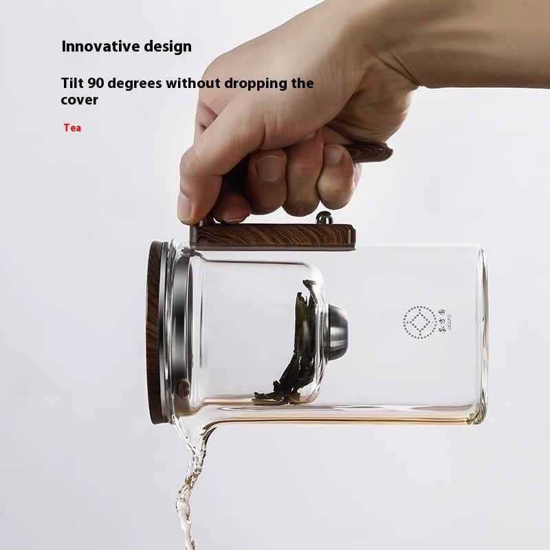 Glass teapot with wooden handle being tilted to pour tea, on a light gray background.